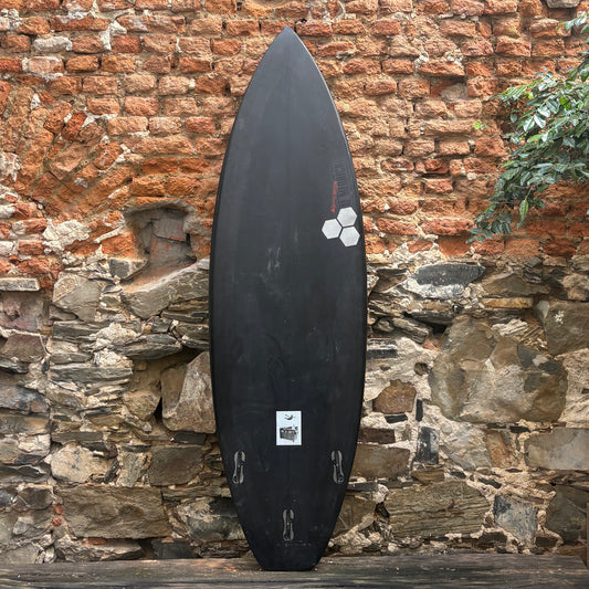 A black surfboard standing upright against a rustic brick and stone wall, with a small white sticker near the bottom and a white geometric logo near the upper middle.