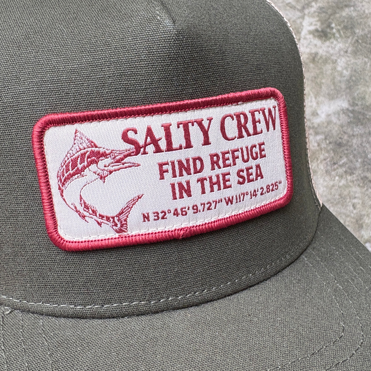 Close-up of a dark olive green trucker hat featuring a rectangular embroidered patch with a red border, red text reading 'SALTY CREW FIND REFUGE IN THE SEA' and a red fish illustration on a white background.