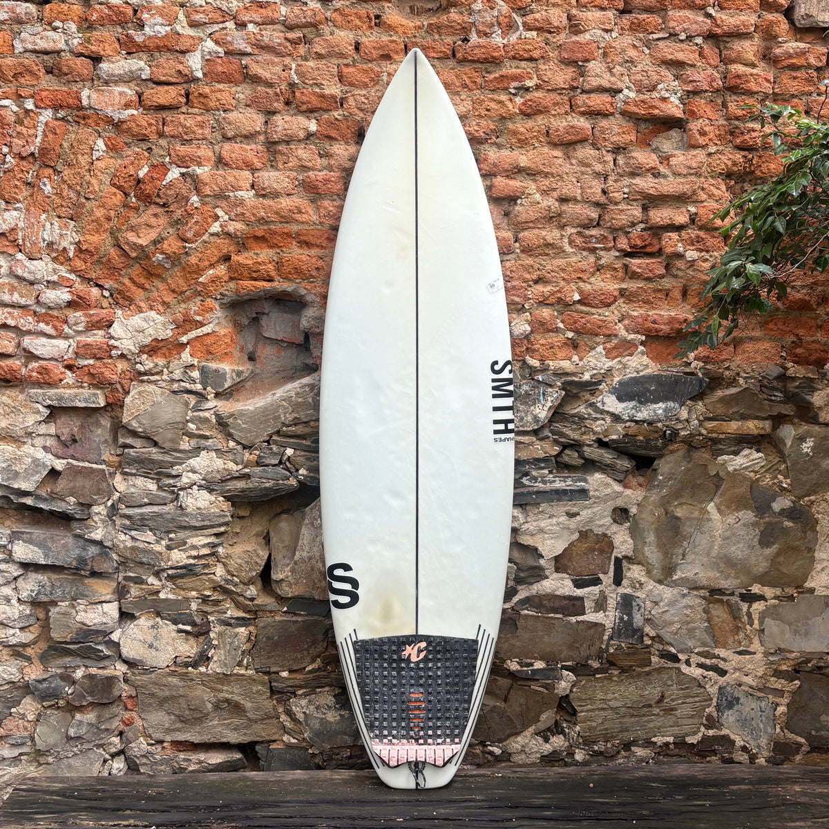 A white surfboard with black graphics and a black footpad, leaning against a rustic brick and stone wall.