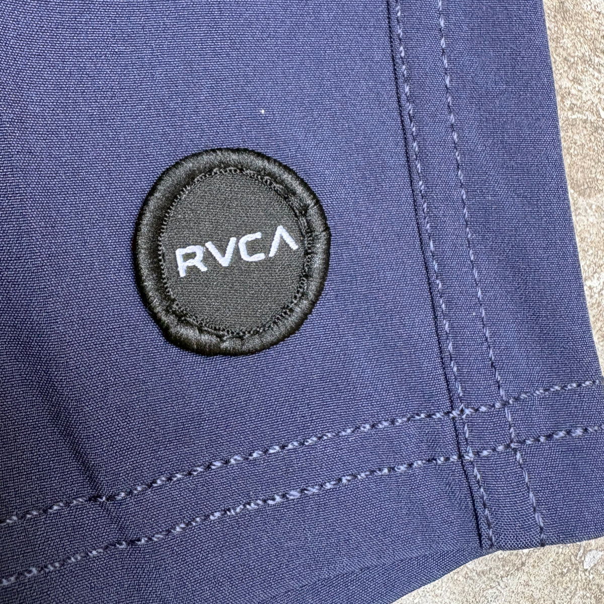 Close-up of a navy blue fabric item with a circular black embroidered patch featuring the white RVCA logo.
