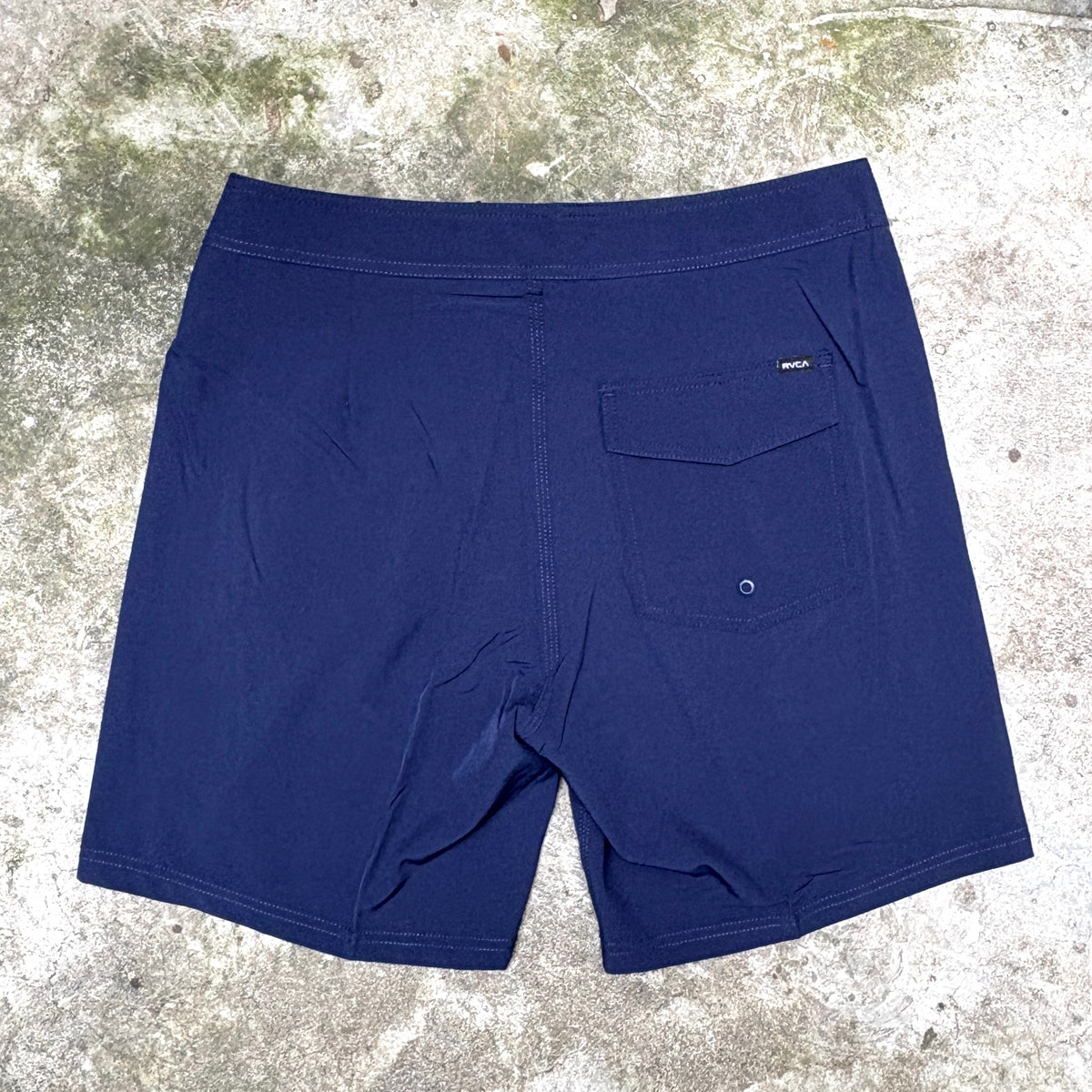 A pair of navy blue board shorts laid flat on a textured concrete surface, showing the back pocket and waistband.