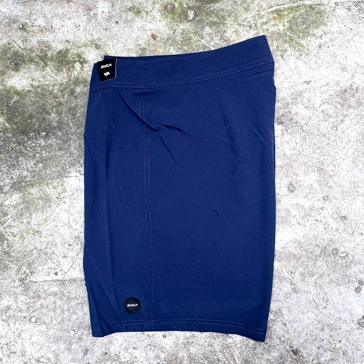 A pair of navy blue board shorts laid flat on a textured concrete surface. The shorts feature a black brand patch on the waistband and a small black circular logo near the lower hem.