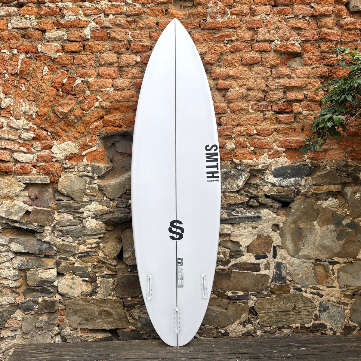 A white surfboard with black graphics and the brand name 'SMITH' leaning against a rustic brick and stone wall.