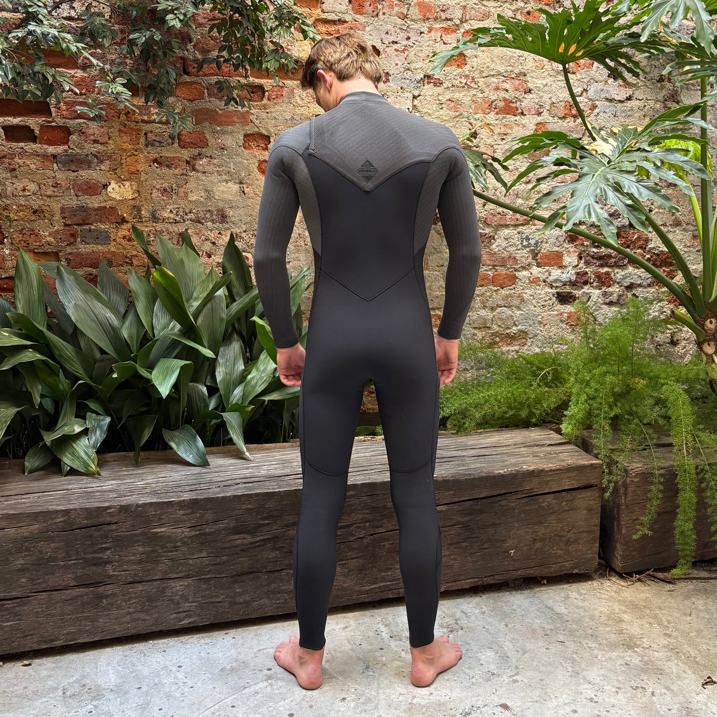 Person wearing a black wetsuit standing in front of a brick wall with plants.