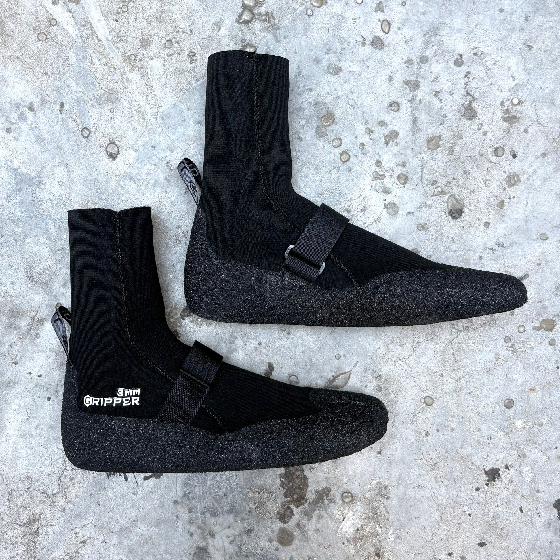 Pair of black neoprene boots with buckles on a textured concrete surface