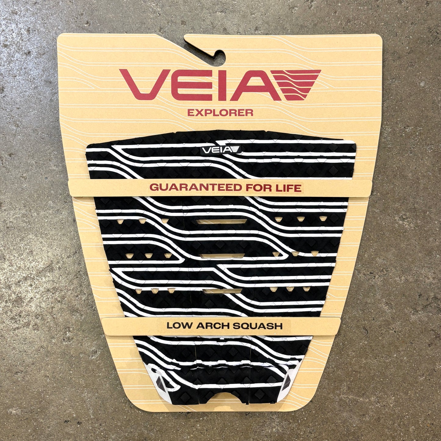 A Veia Explorer low arch squash paddle grip pad in black and white wavy pattern, mounted on its original beige cardboard backing against a gray concrete floor.