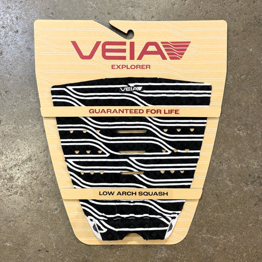 A Veia Explorer low arch squash paddle grip pad in black and white wavy pattern, mounted on its original beige cardboard backing against a gray concrete floor.