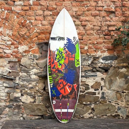 Colorful JS INDUSTRIES surfboard with abstract design against a rustic stone wall.