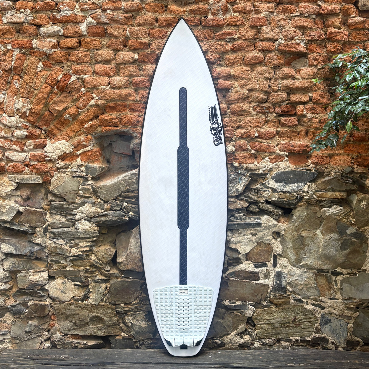 A white surfboard with a black center stripe and black graphic logo near the tail, leaning against a rustic brick and stone wall.