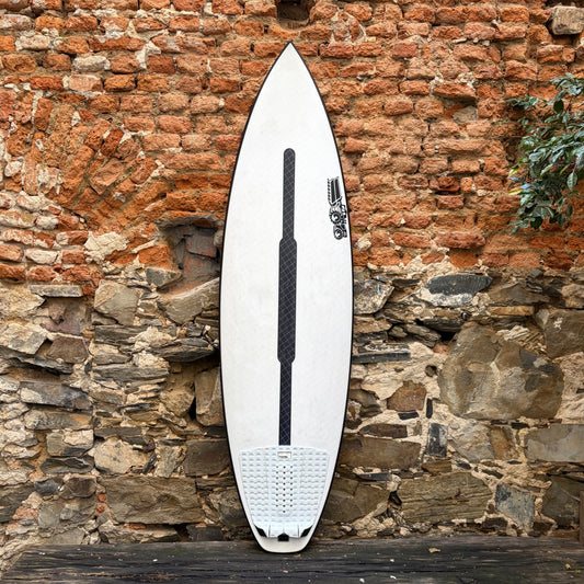 A white surfboard with black grip tape and a black leash attached, leaning against a rustic brick and stone wall.