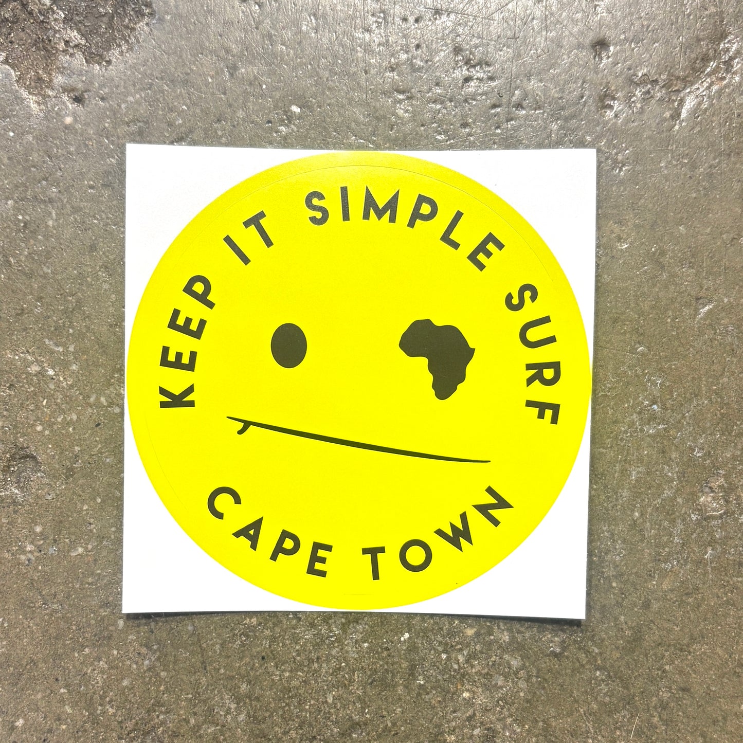 A square white sticker featuring a bright yellow circle with the text 'KEEP IT SIMPLE SURF' and 'CAPE TOWN' in black, plus a minimalist face with one black dot and one black silhouette, placed on a textured gray concrete surface.