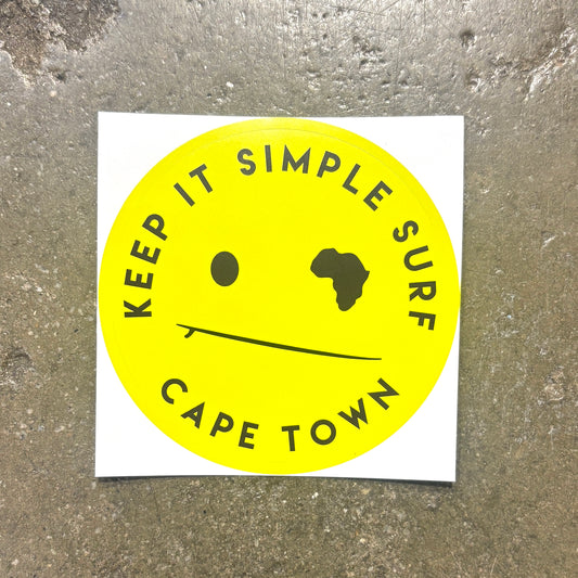 A square white sticker featuring a bright yellow circle with the text 'KEEP IT SIMPLE SURF' and 'CAPE TOWN' in black, plus a minimalist face with one black dot and one black silhouette, placed on a textured gray concrete surface.
