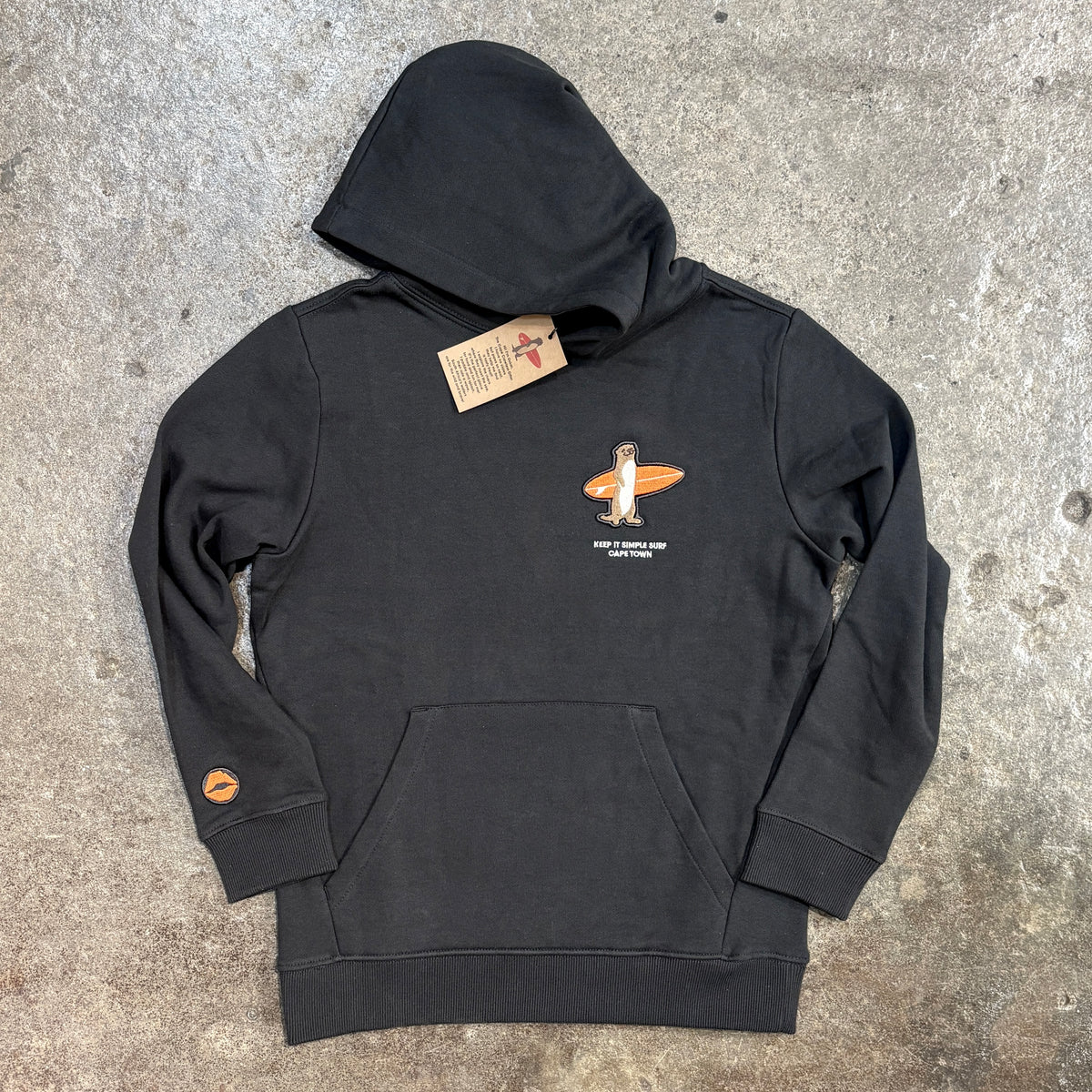 A black pullover hoodie laid flat on a textured concrete surface, featuring a small orange and white graphic on the left chest with the text 'JACKIE DOUGLAS WALKER SURFER' and a small orange circular patch on the right sleeve.