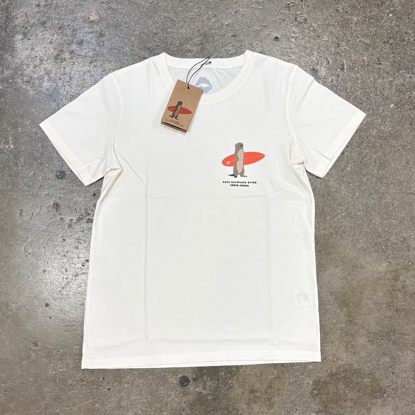 White Kiss surf Store t-shirt with a graphic of a Otter on a surfboard and a tag, on a textured gray surface.