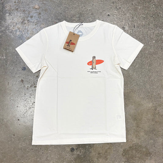 White Kiss surf Store t-shirt with a graphic of a Otter on a surfboard and a tag, on a textured gray surface.