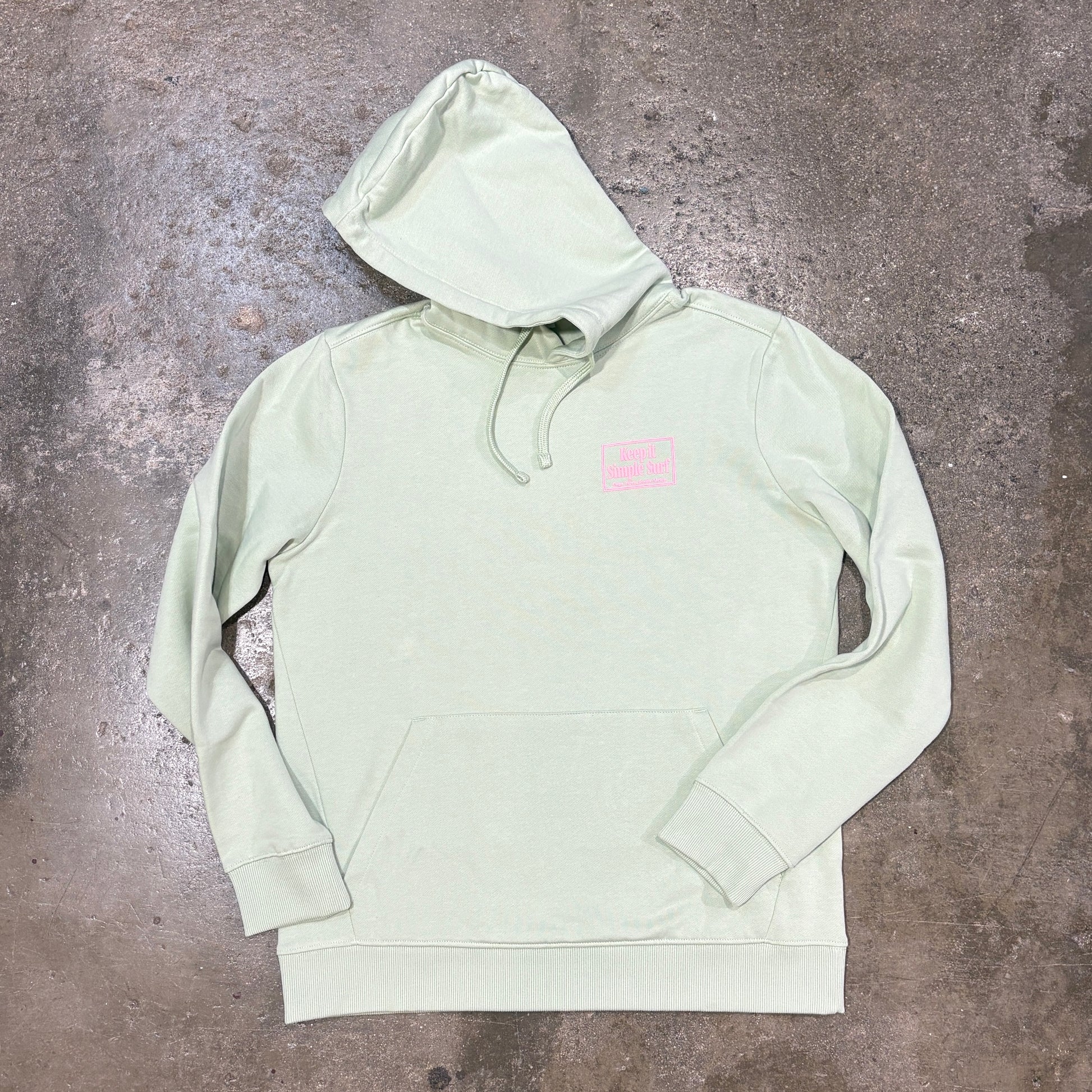 A light sage green pullover hoodie with a small pink rectangular logo patch on the left chest, laid flat on a textured gray concrete floor.