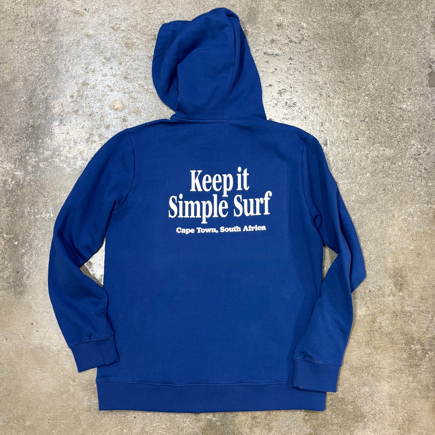 A royal blue hoodie laid flat on a textured concrete surface, featuring the white printed text 'Keep it Simple Surf' and 'Cape Town, South Africa' on the back.