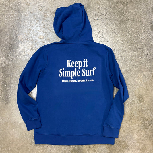 A royal blue hoodie laid flat on a textured concrete surface, featuring the white printed text 'Keep it Simple Surf' and 'Cape Town, South Africa' on the back.