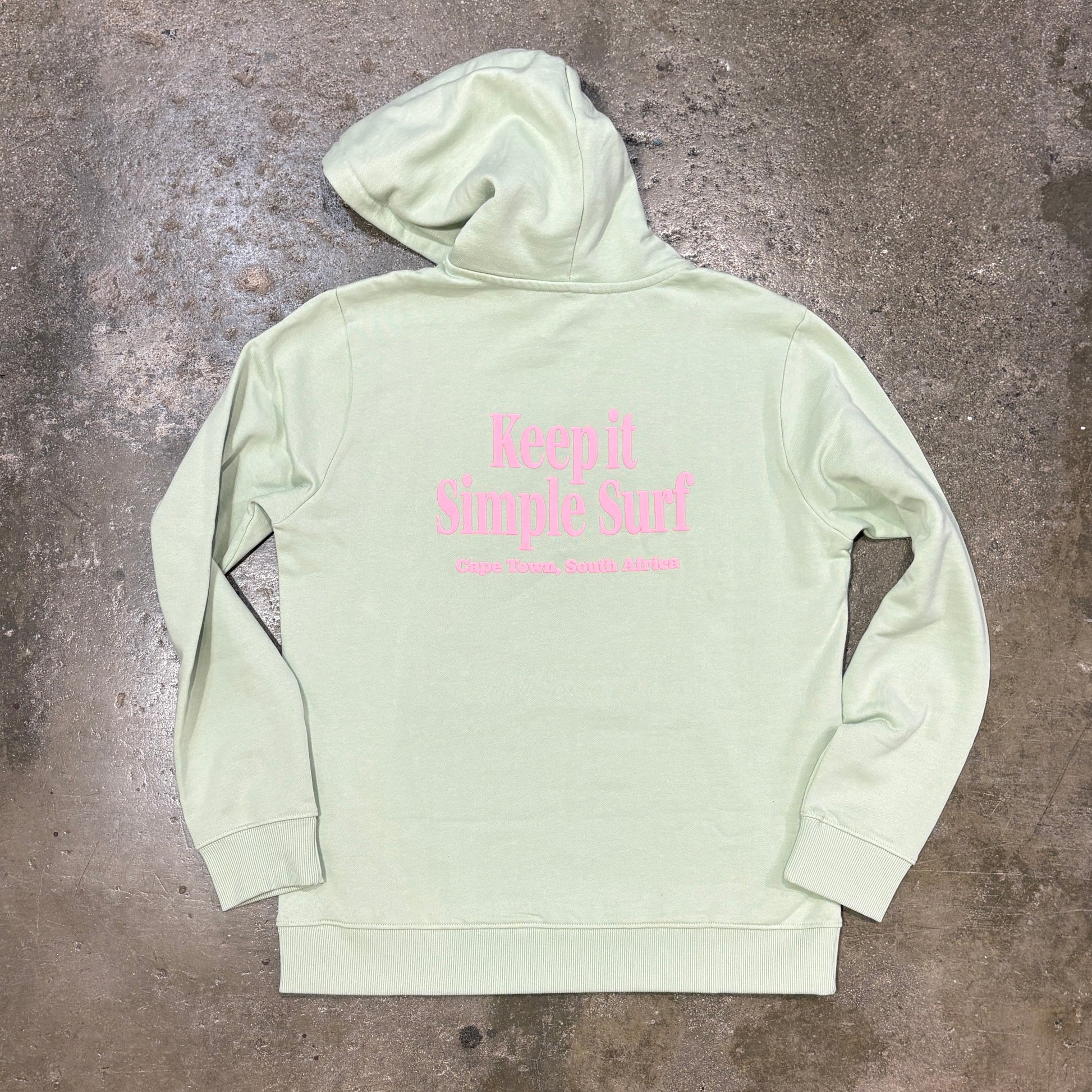 A light sage green hoodie with pink text reading 'Keep It Simple Surf' on the back, laid flat on a textured concrete floor.