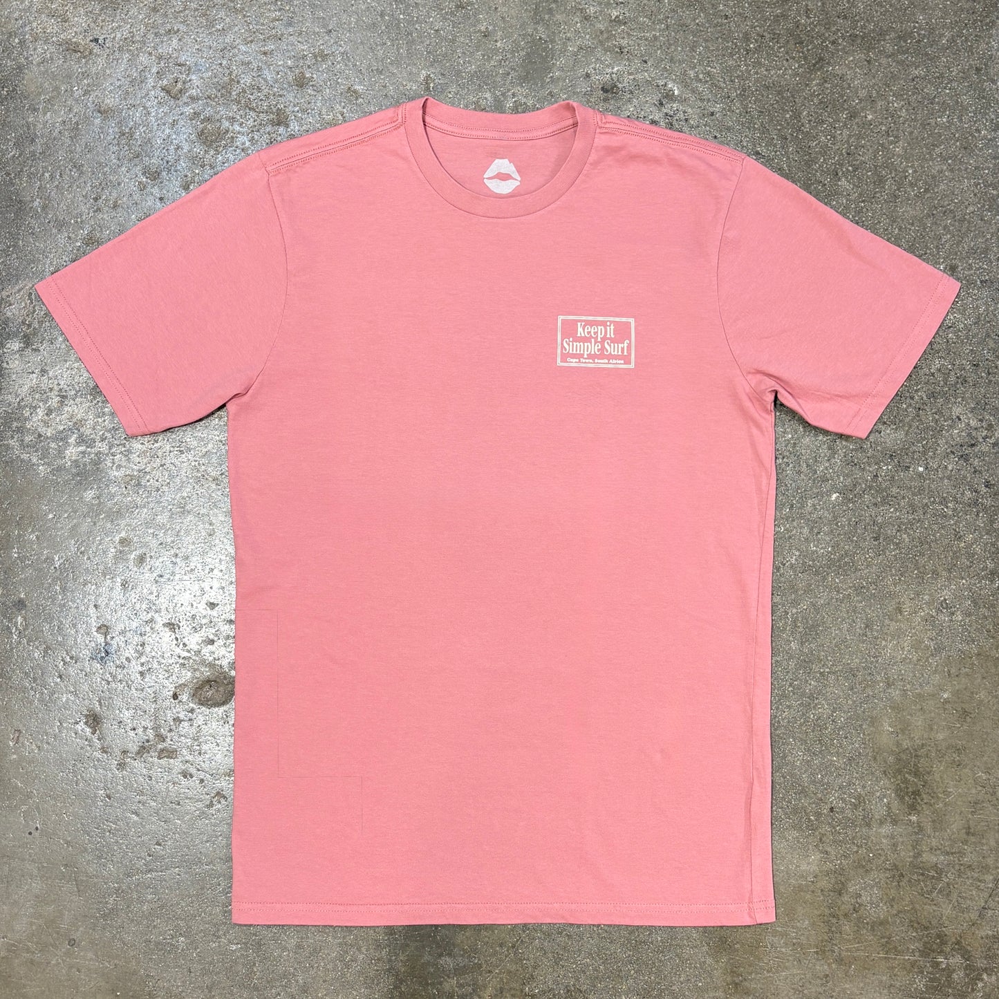 A pink short-sleeve crewneck t-shirt with a small white graphic on the left chest reading 'keep it simple surf . Cape Town, South Africa' laid flat on a textured gray concrete surface.
