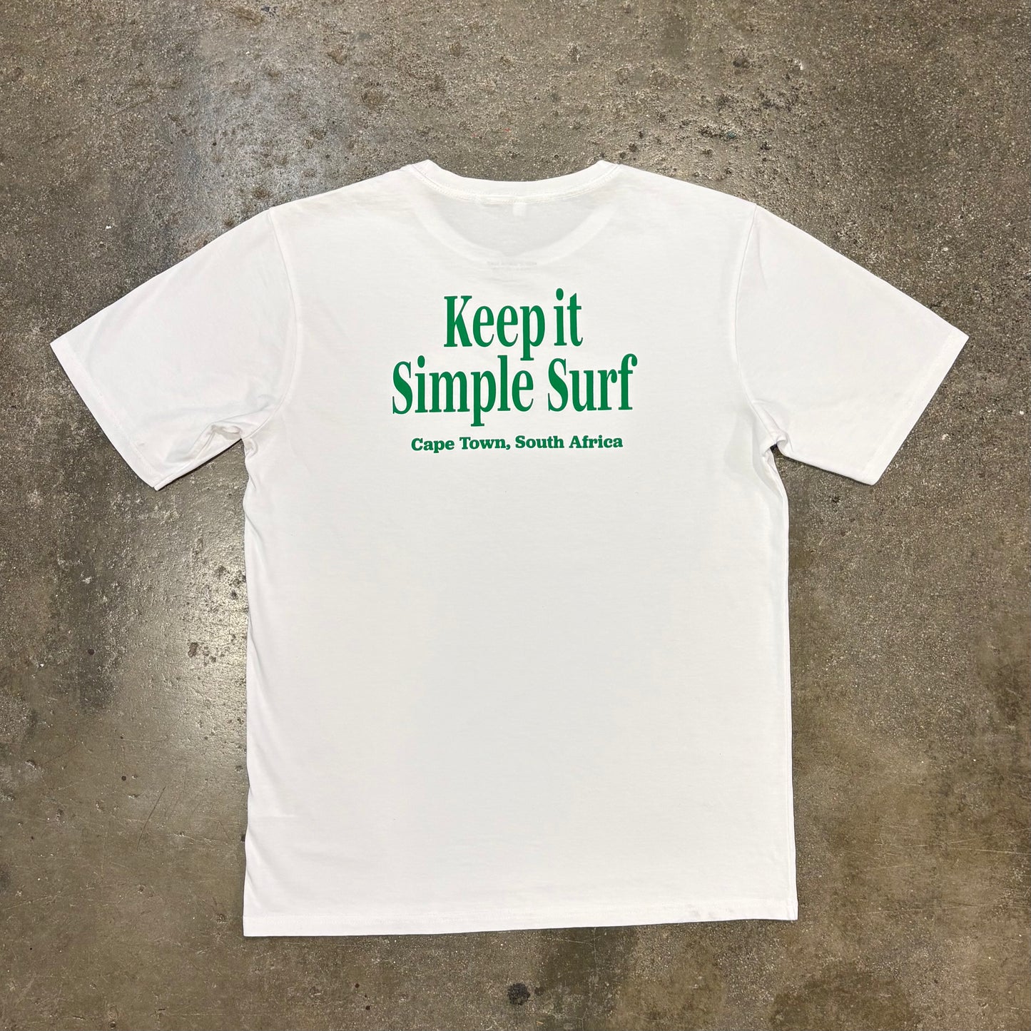 White t-shirt with green text 'Keep it Simple Surf Cape Town, South Africa' on a concrete surface