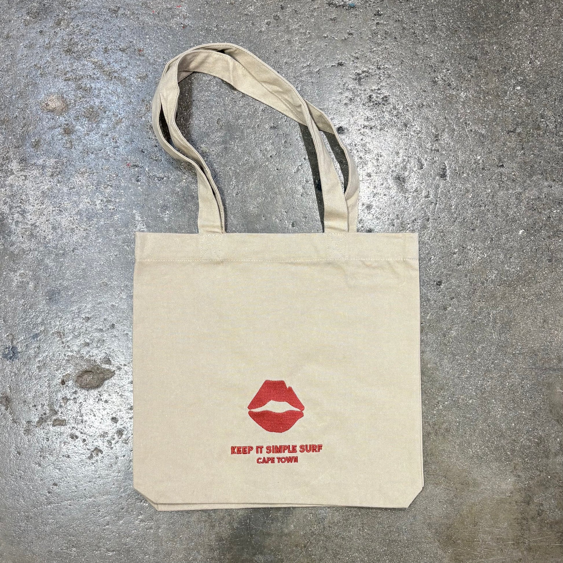 Beige tote bag with red lips graphic and text on a textured gray surface