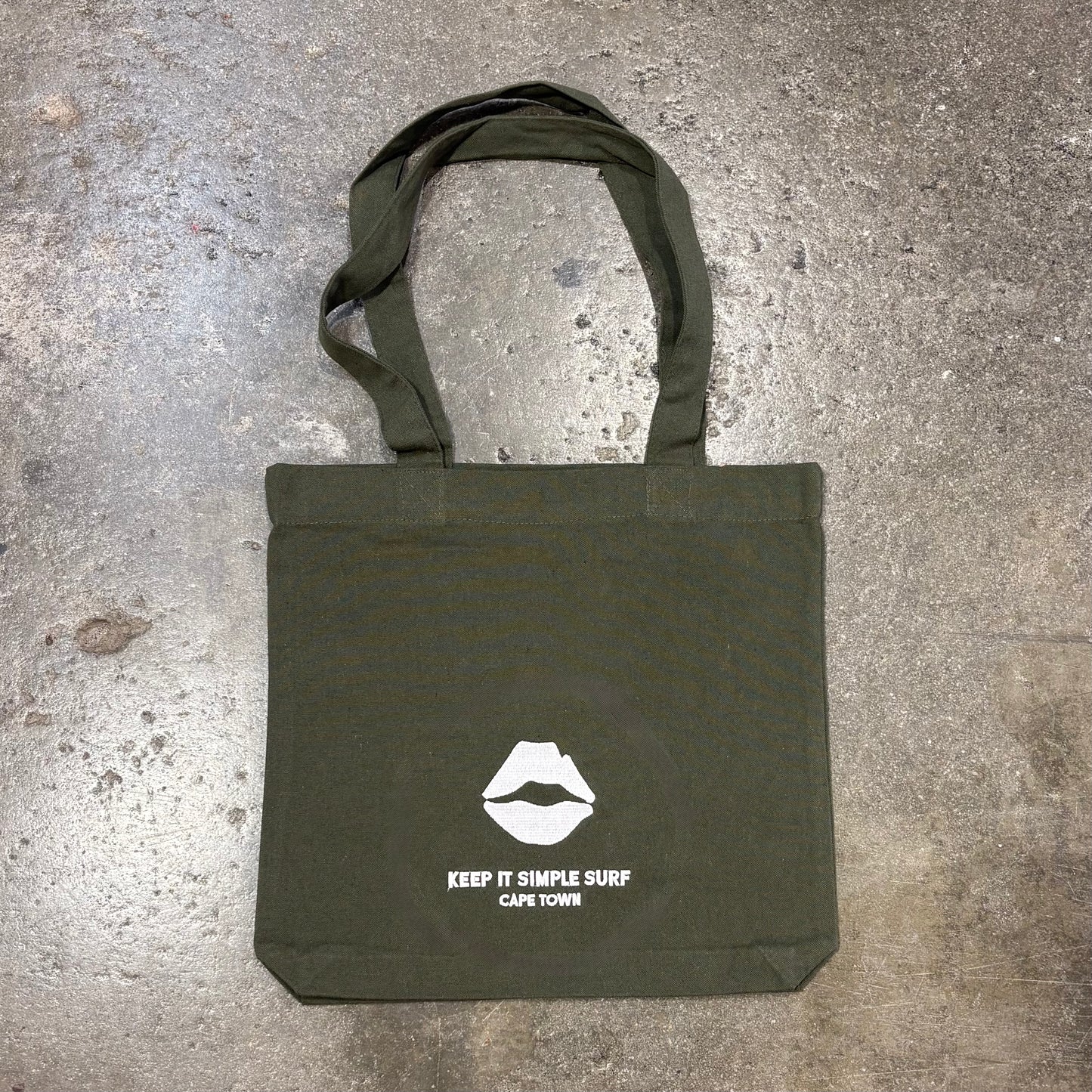 Green Kiss Surf Store  tote bag with white logo and text on a concrete surface