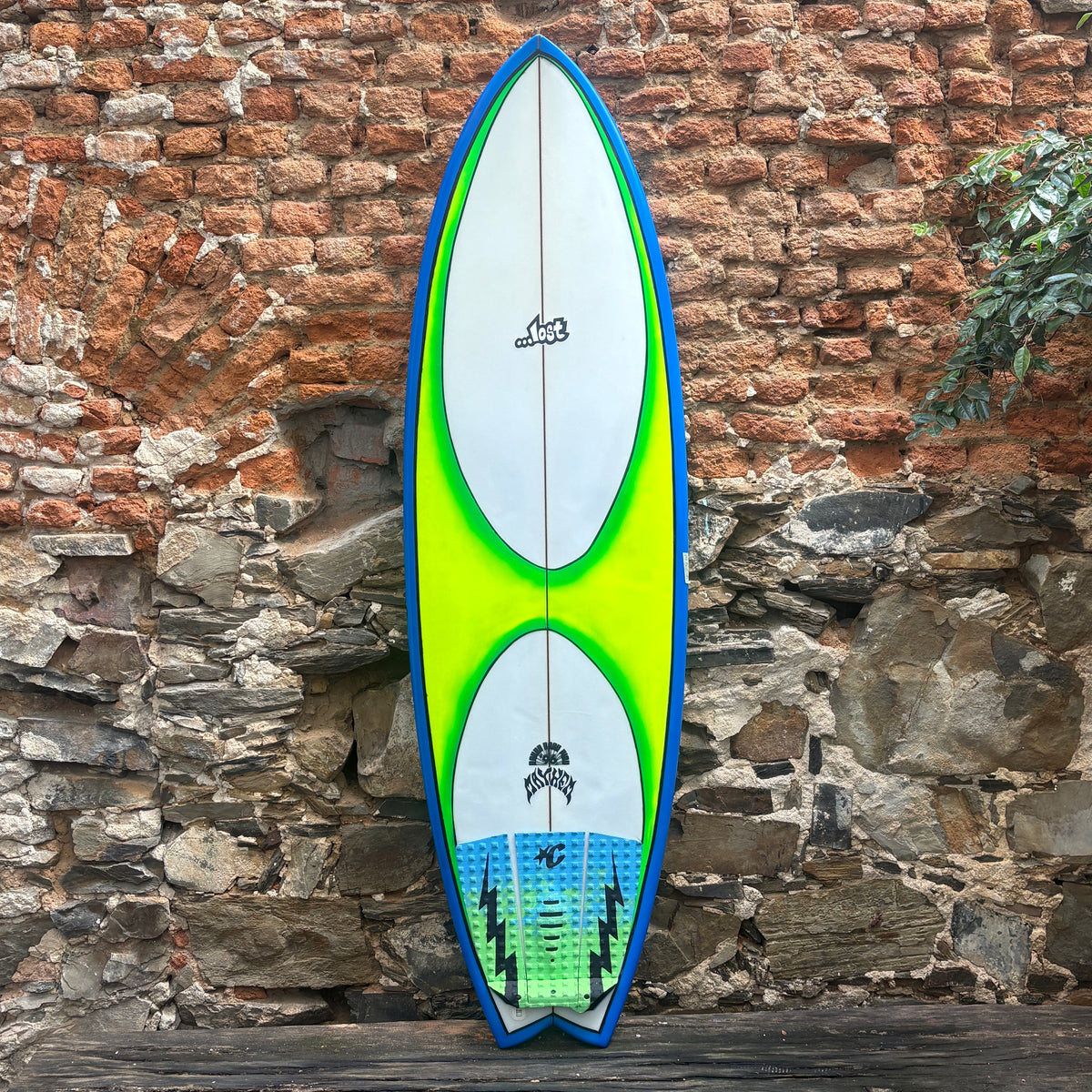 A long surfboard with a blue border, white center, bright neon green graphic, and blue-green tail section, leaning against a rustic brick and stone wall.
