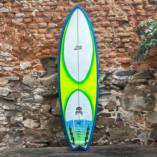 A long surfboard with a blue border, white center, bright neon green graphic, and blue-green tail section, leaning against a rustic brick and stone wall.