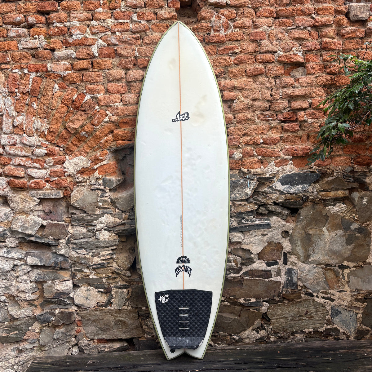 A white surfboard with black graphics and a black traction pad, leaning against a rustic brick and stone wall.