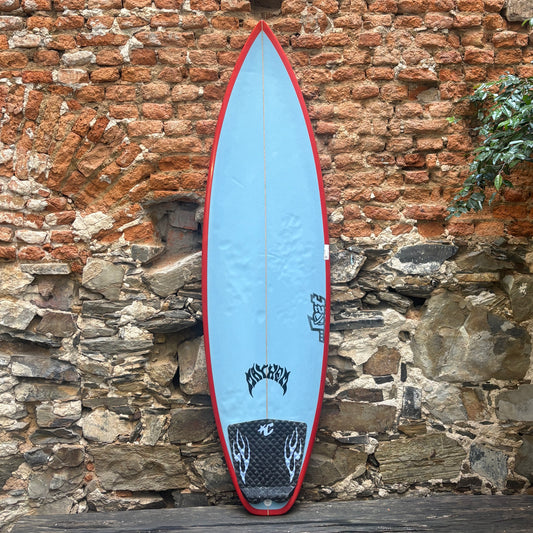 A light blue surfboard with a red border, standing upright against a rustic stone and brick wall. The board features black graphics and the brand name 'Mossy Oak' near the tail.