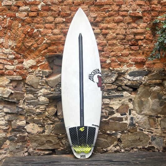 A white surfboard with black and red graphics, including the 'Kings of NY' logo, leaning against a rustic brick and stone wall.