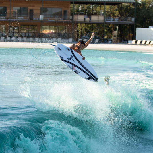 A surfer in mid-air performing a jump on a white surfboard with black graphics, set against a turquoise pool wave with a resort building in the background.