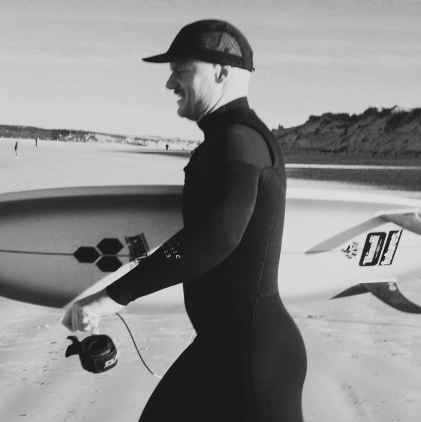 Men in Manera wetsuit carrying surfboard on beach