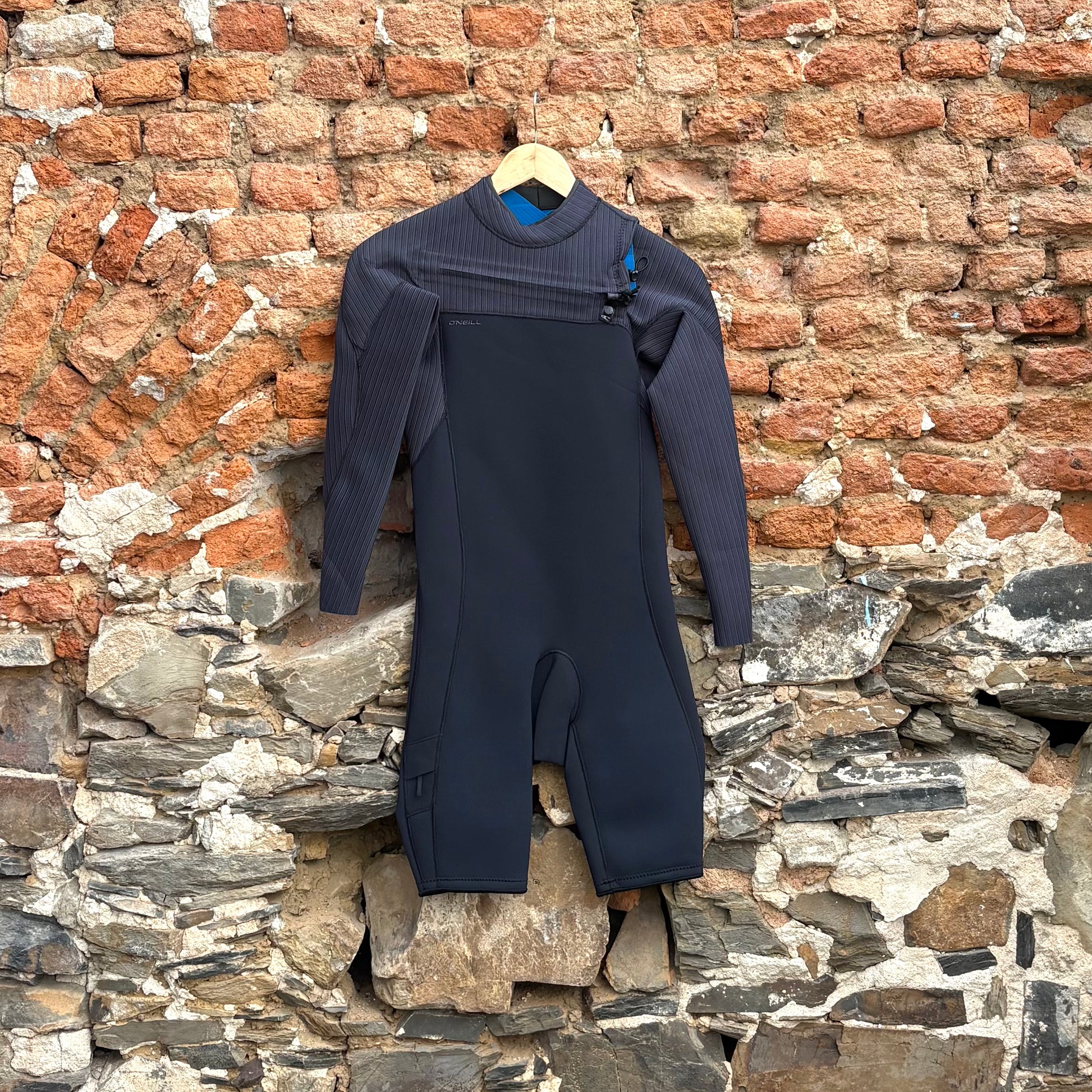 Black Shortie wetsuit on a hanger against a rustic brick wall.