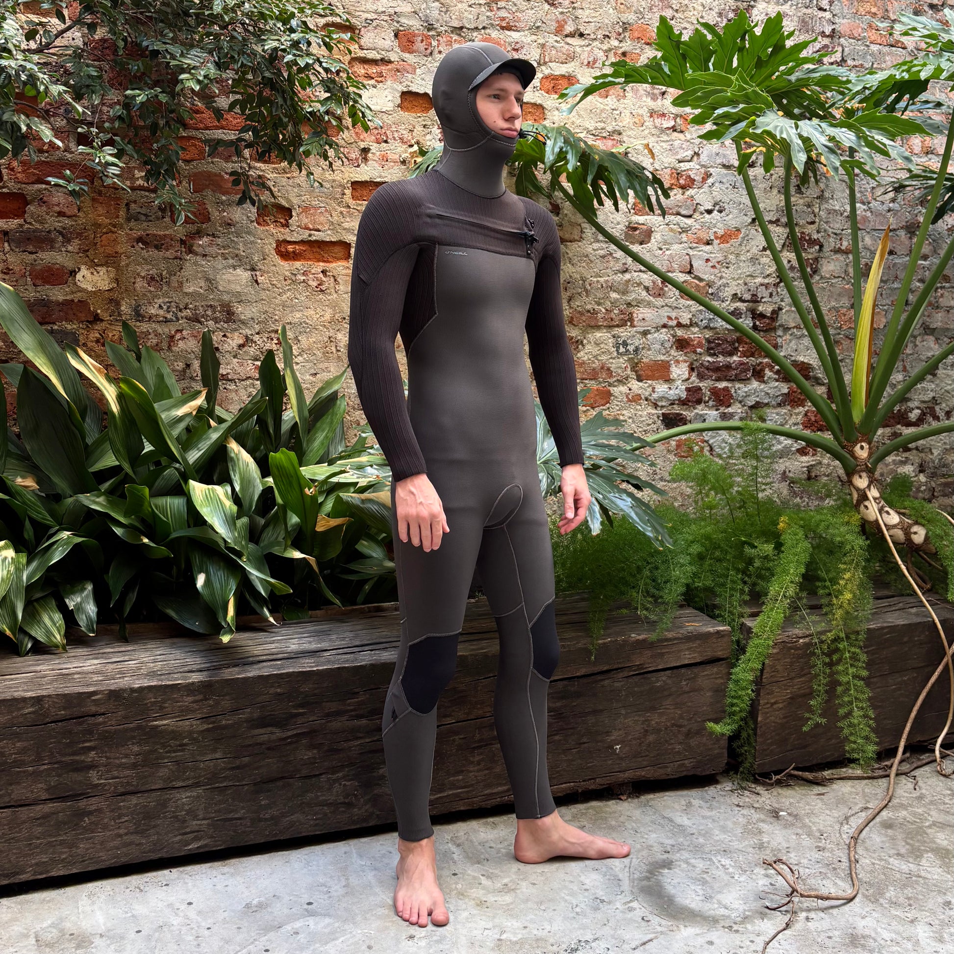 Person wearing a full-body wetsuit standing outdoors with plants and a brick wall in the background