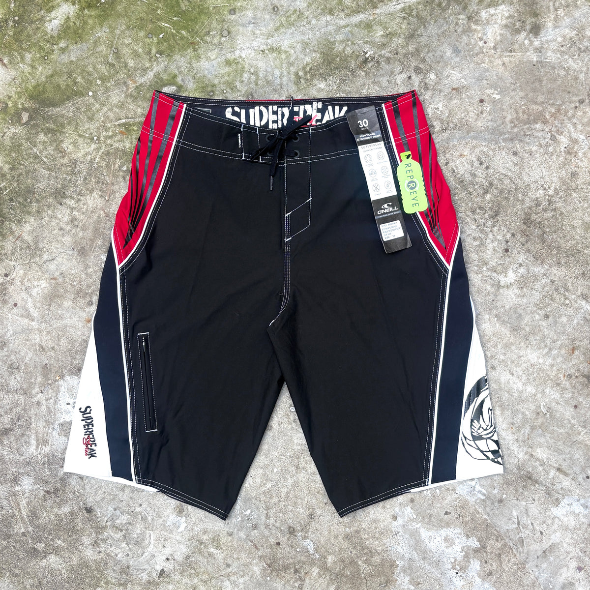 Black board shorts with red and white side panels, laid flat on a gray concrete surface. The shorts feature the brand name 'SUPERFREAK' printed on the waistband, a drawstring waist, and hangtag tags attached near the right side.
