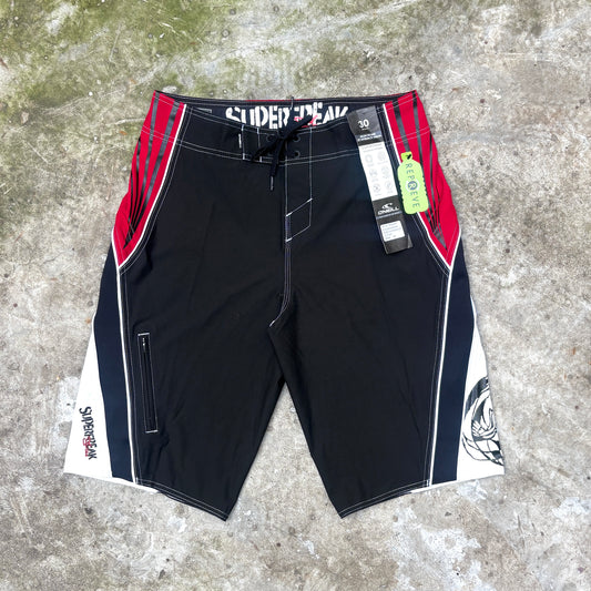 Black board shorts with red and white side panels, laid flat on a gray concrete surface. The shorts feature the brand name 'SUPERFREAK' printed on the waistband, a drawstring waist, and hangtag tags attached near the right side.