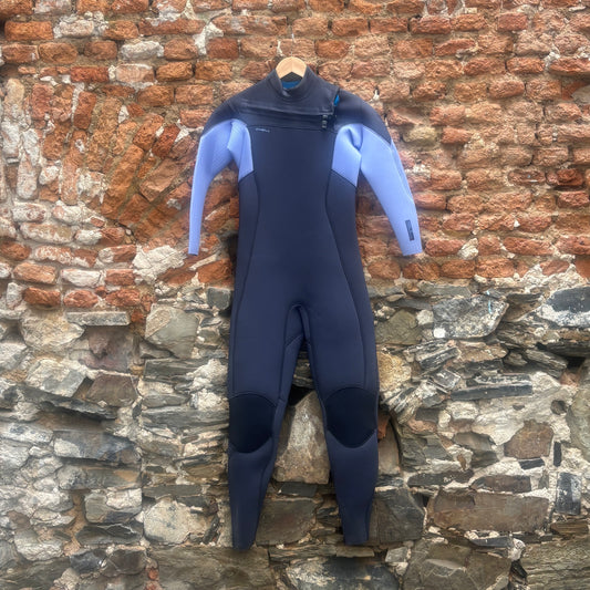 A full-body wetsuit hanging on a wooden hanger against a rustic brick and stone wall. The wetsuit is dark navy blue with light blue panels on the shoulders and sleeves.