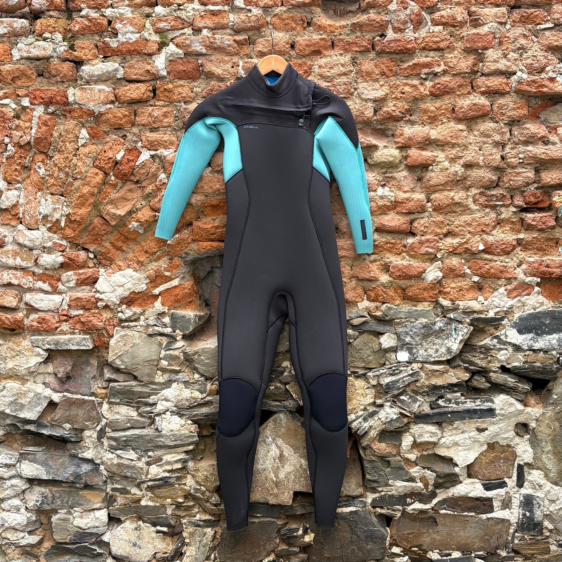 Black and teal O'Neill Hyperfreak wetsuit hanging on a brick wall.