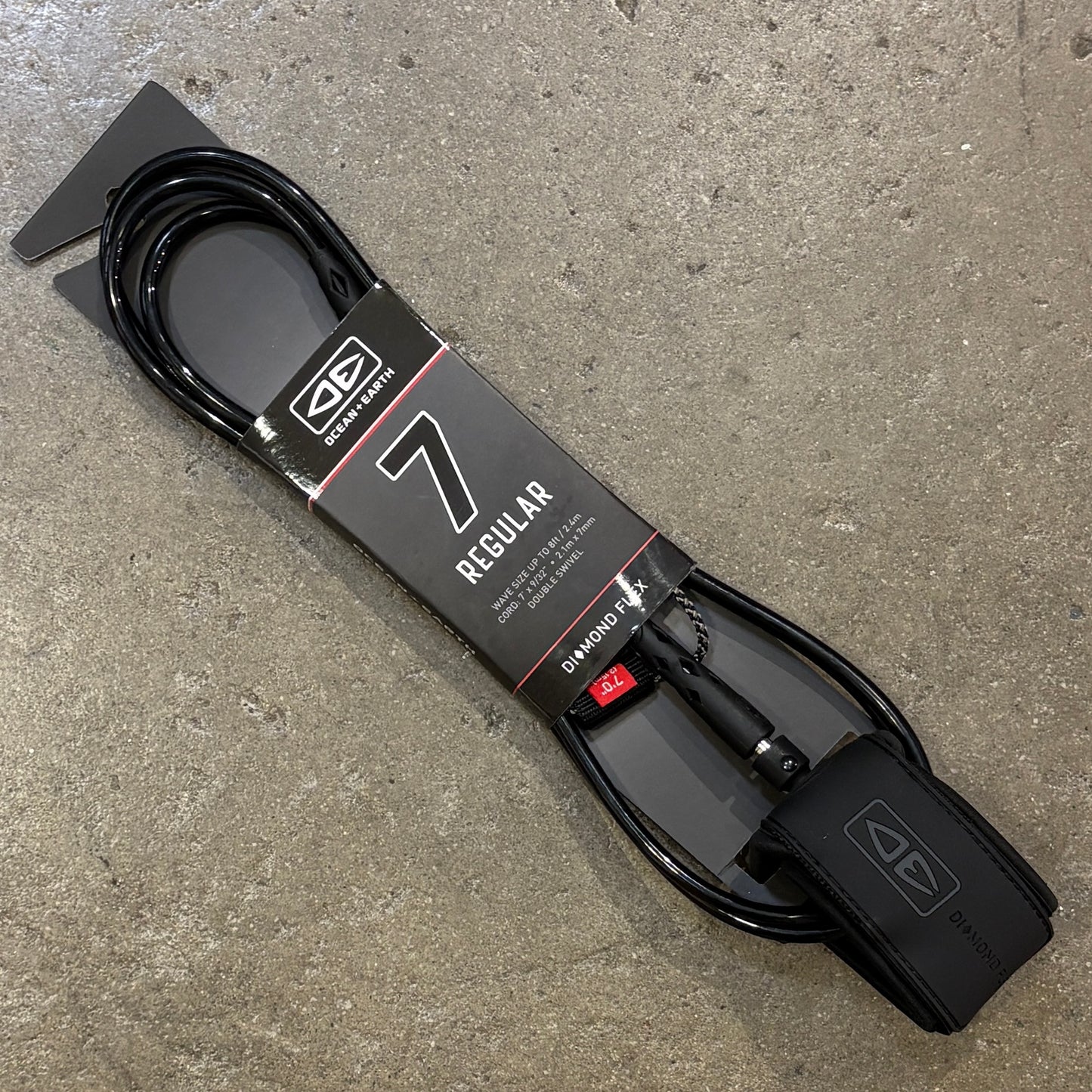 A black  7 Regular surf leash with its retail packaging, placed diagonally on a textured gray concrete floor.