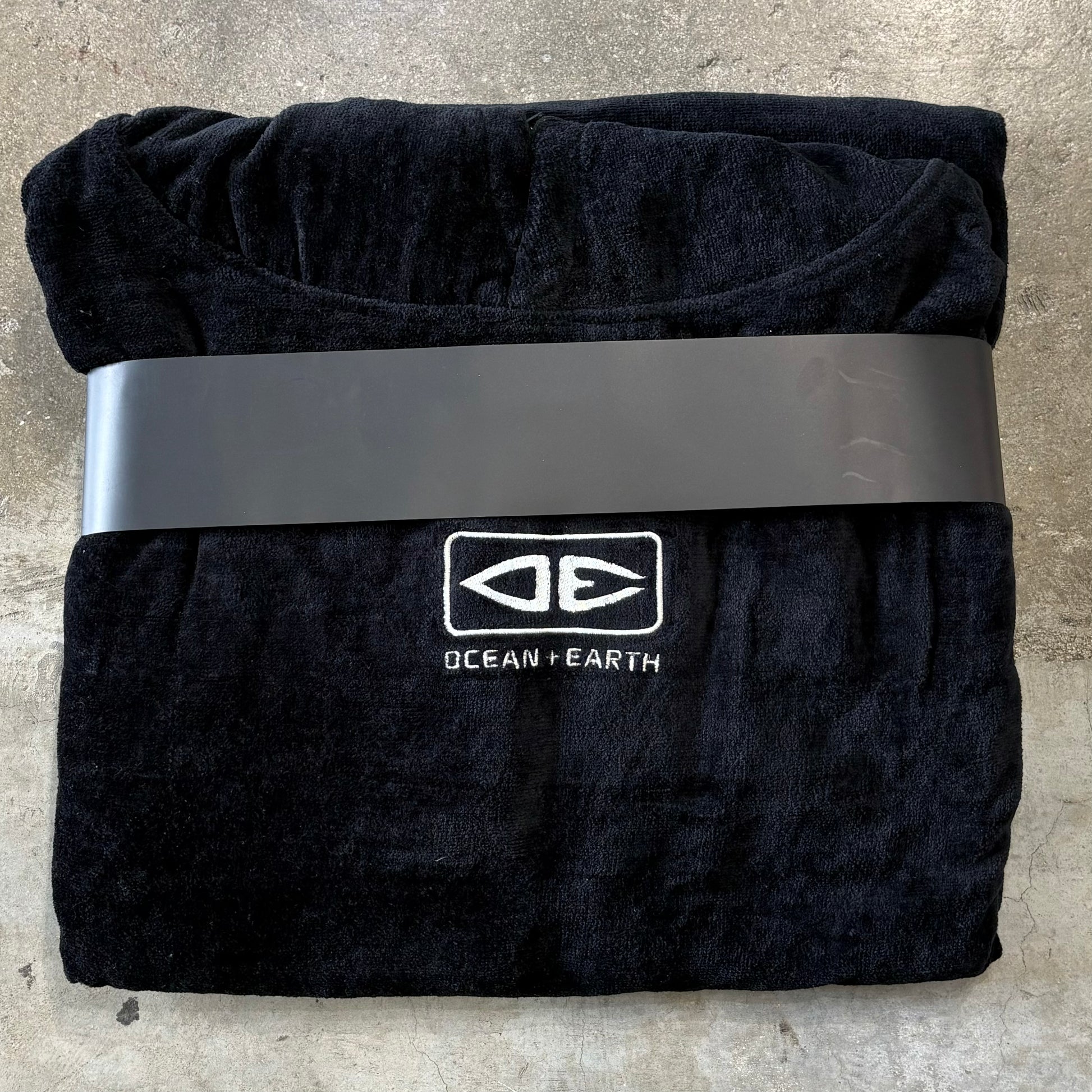 Folded Surf Towel in black with Ocean& Earth Logo on a concrete floor