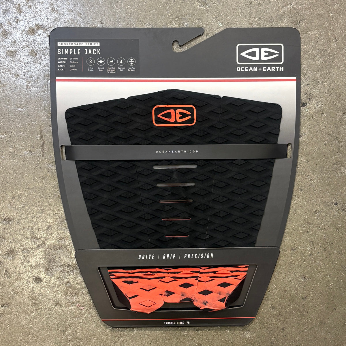 Ocean + Earth Simple Jack traction pad in black with orange accents, shown in its retail blister packaging against a concrete floor background.