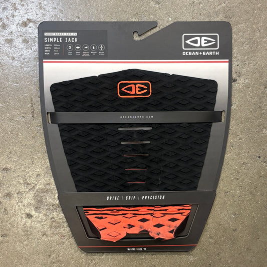 Ocean + Earth Simple Jack traction pad in black with orange accents, shown in its retail blister packaging against a concrete floor background.
