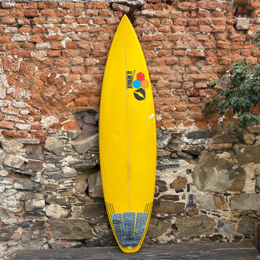 Yellow surfboard with a colorful logo against a rustic stone wall.