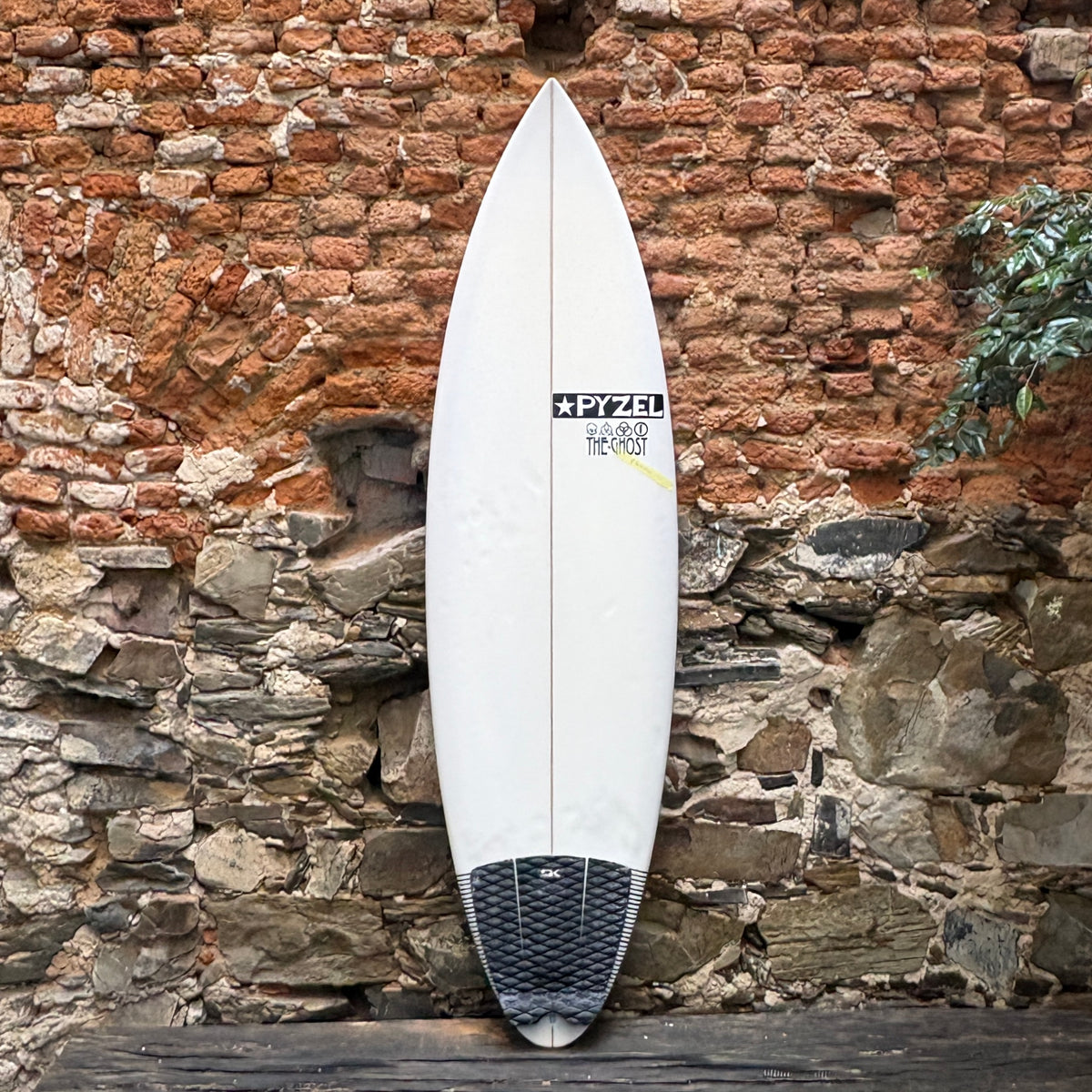 A white surfboard with black and yellow graphics and a black grip pad at the tail, leaning against a rustic brick and stone wall.