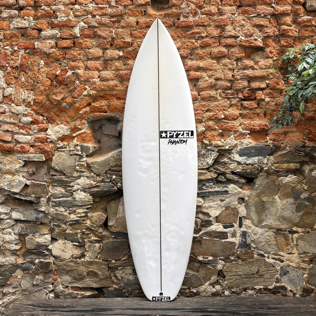 A white surfboard with black Zetl branding, leaning vertically against a rustic brick and stone wall.