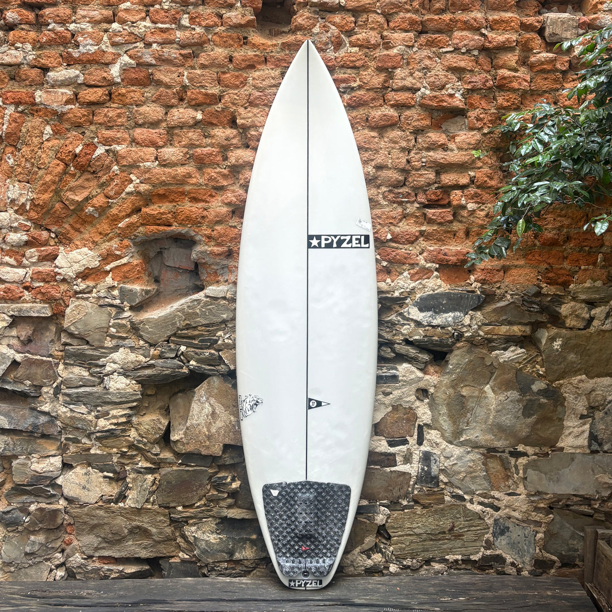 A white Pyzel surfboard with a black grip pad and black logo, leaning against a rustic brick and stone wall.