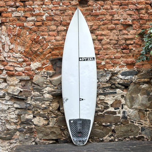 A white surfboard with a black Pyzel logo and a black grip tape patch near the tail, leaning against a rustic red and gray brick wall.