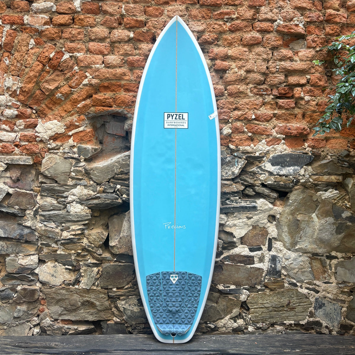 A light blue Pyzel surfboard with a dark gray traction pad, leaning against a rustic brick and stone wall.